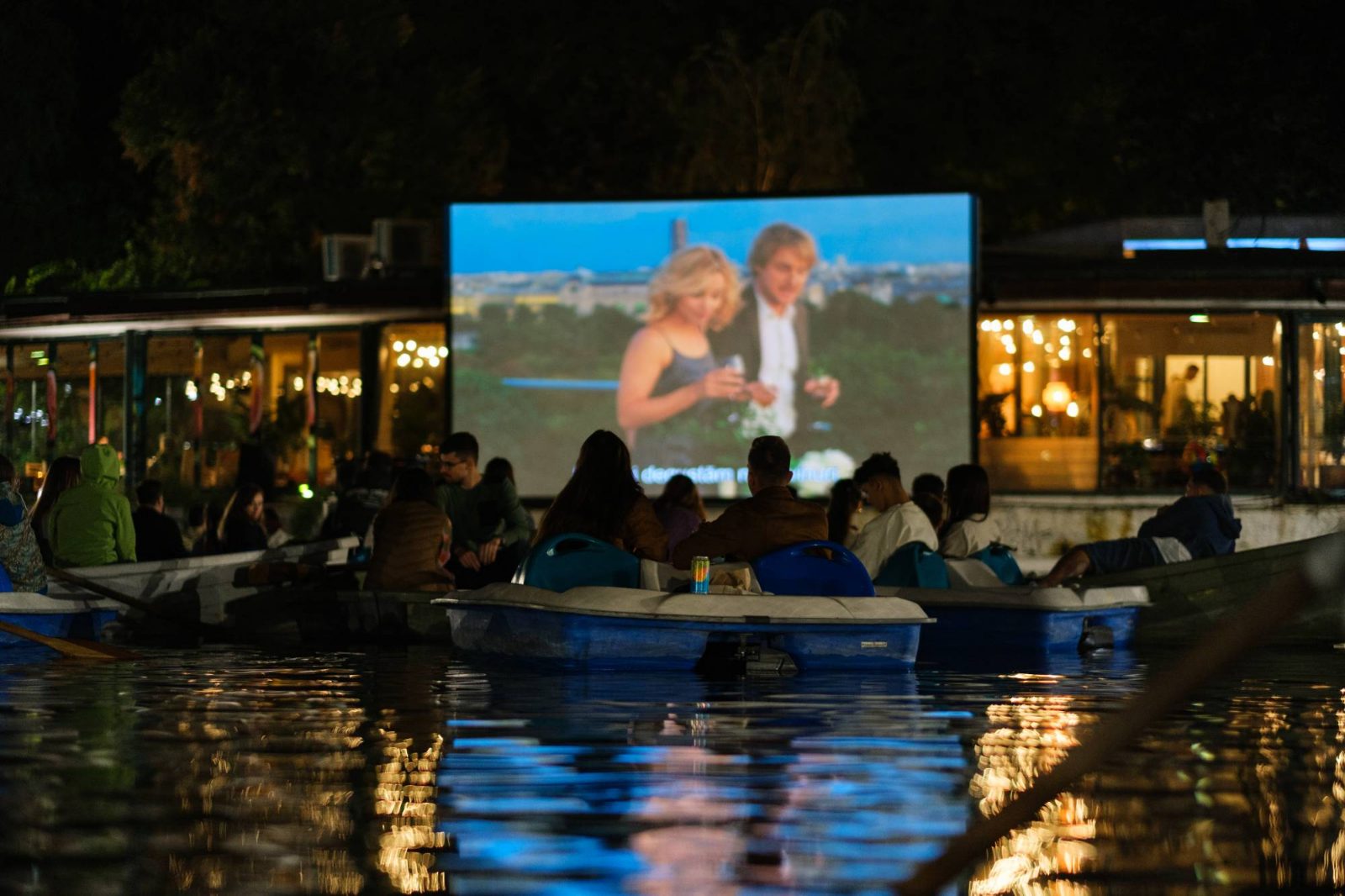 Public în bărci pe lac, în fața ecranului de cinema, seara.