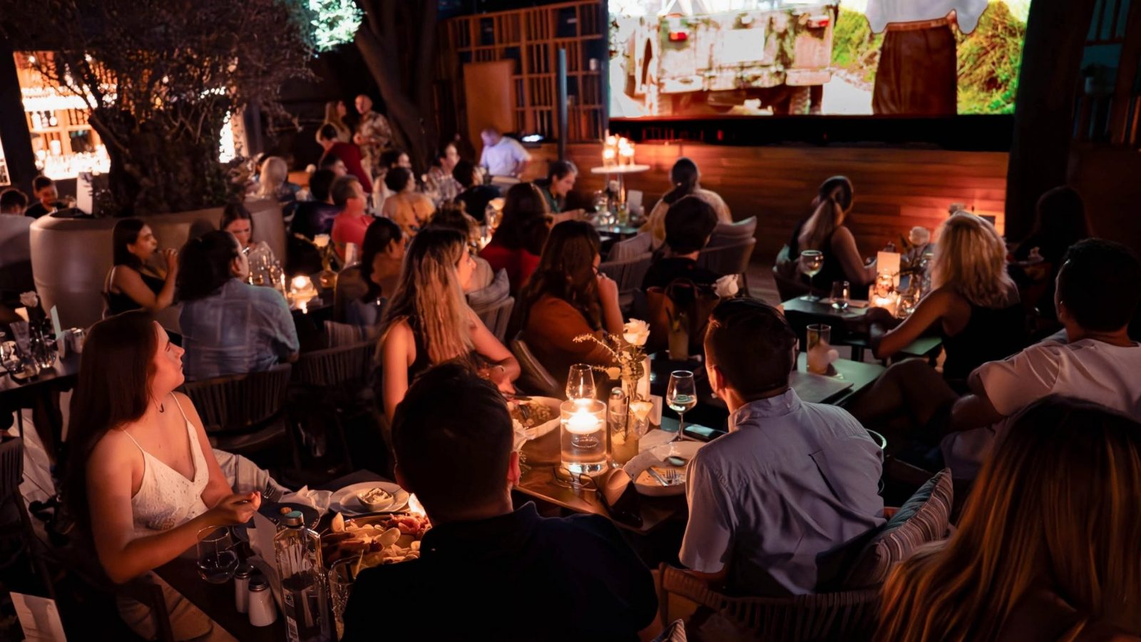 Audience seated at tables on the terrace, in front of the cinema screen.