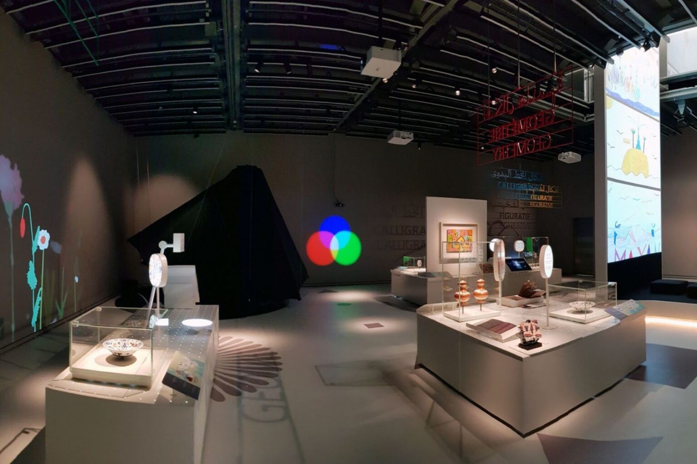 Vedere largă a expoziției, cu vitrine, proiecții și forme colorate