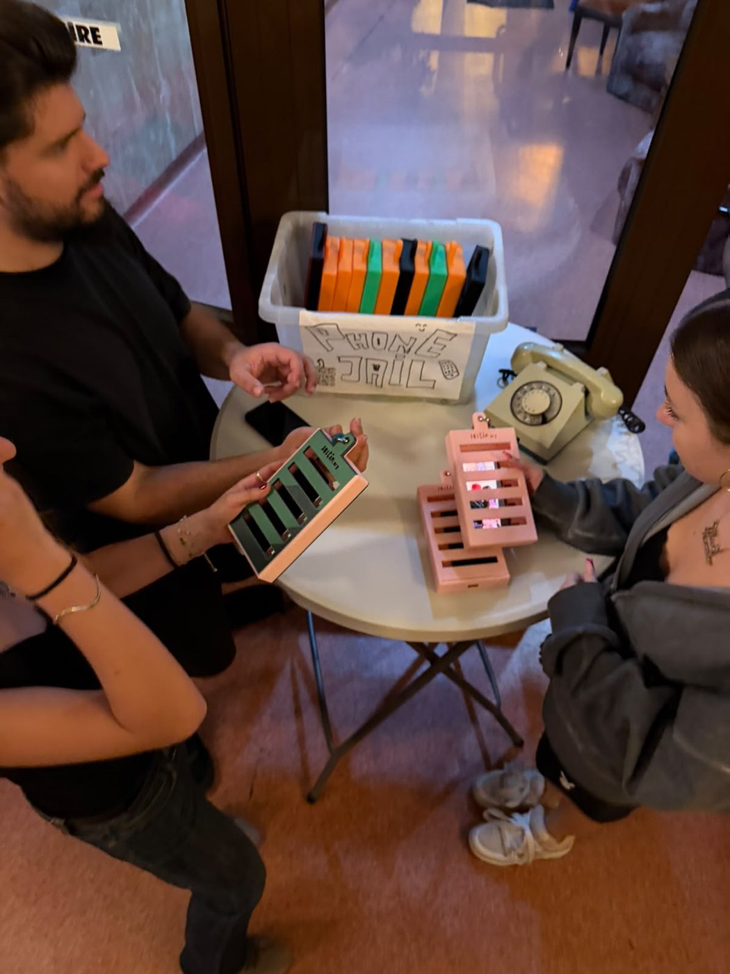 Cutia „Phone Jail” plină cu carcase colorate, pe masa de la intrarea în sală.